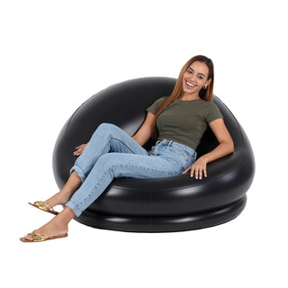 A person with long hair, dressed in a green T-shirt, blue jeans, and sandals, is sitting on a large Pool Candy 8-Ball Black & White Inflatable Chair. They are smiling and leaning back comfortably, with their legs casually crossed at the ankles against a plain white background.