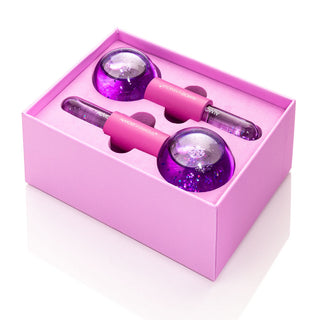 A pink box from Almost Famous contains two purple, globe-shaped Cryo Facial Treatment Massage Globes with attached handles, resting in designated slots. The objects have a shiny, glittery texture. The open box reveals the neatly arranged massage globes, perfect for enhancing your skincare routine and elevating your facial experience. Enjoy this FREE GIFT 🎁!