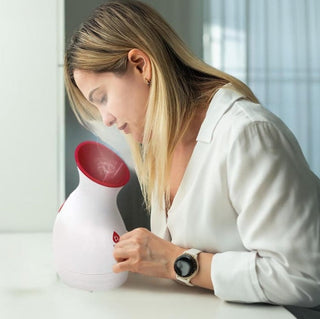 A blonde-haired woman sits at a table, wearing a white blouse and wristwatch, using the Pursonic Pore Purifying Facial Steamer with its distinctive red nozzle. Leaning over the device, she enjoys a moisturizing spa treatment as steam envelops her face. The softly blurred background, adorned with light curtains, adds to the serene atmosphere.
