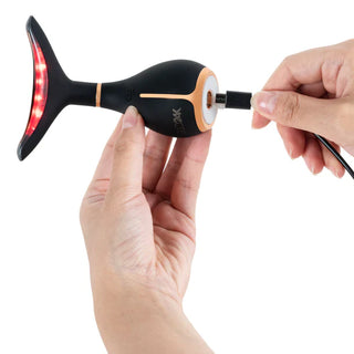 Hands are shown holding a black and orange fish-shaped electronic device with a tail that has red LED lights. One hand is holding the GLO24K Skin Rejuvenation LED Beauty Device (for Face & Neck) while the other is inserting a charging cable into its port, highlighting its advanced age-defying benefits and LED Light Therapy Technology.