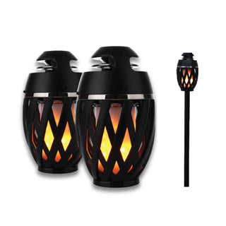 Image shows two black Gabba Goods Bluetooth LED Flame Speakers with crisscrossing lattice patterns on their cylindrical bodies, both emitting a warm, orange glow. One torch is also shown mounted on a black stake, indicating it can be used as a yard or garden decoration with Bluetooth Flame Speaker functionality for added ambiance.