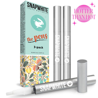 Image of Snapwhite Advanced Teeth Whitening Travel Pens (3-Pack), featuring three silver pens labeled "Snapwhite" filled with a gentle gel formula. The pastel-colored box boasts floral designs, the text "the pens," and a smiling tooth logo. A pink circular emblem with the text "HOTTER THAN HOT" decorates the top right corner.