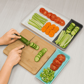 A person uses the Madison York Mess-Free "Cut & Carry" Kitchen Prep Station to slice a cucumber on its bamboo cutting board with trays. Three rectangular plates neatly display various sliced vegetables: celery, carrots, and tomatoes on a white plate; cucumbers and celery on a black plate; sliced tomatoes and diced green peppers on a blue plate. It's truly a mess-free experience.