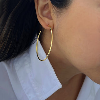 A close-up of a person wearing Sonia Hou Jewelry's Perfect Hoop Earrings. The person has long, dark hair and is dressed in a white collared shirt. The focus is on the large, gold hoop earring and part of the ear, with the rest of the face not fully visible.