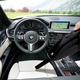 A person uses the Pursonic Handheld Car Vacuum Cleaner (with Adapter Included) featuring 1800PA super suction to clean the interior of a car. The car's dashboard and GPS system are visible, along with the steering wheel and gear shift. The interior is black and white, and there's a scenic landscape visible through the windows.