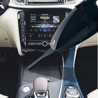 A close-up of a car interior shows a hand using the Pursonic Handheld Car Vacuum Cleaner (with Adapter Included), boasting 1800PA super suction, to clean the center console area. The dashboard screen and control panel in the background display a media interface. The well-maintained, modern car also features a washable reusable filter for added convenience.