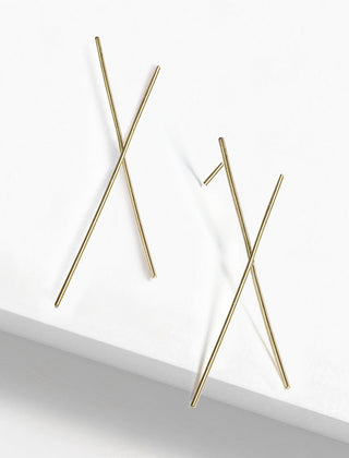 Minimalistic Chopstick Earrings by Sonia Hou Jewelry, composed of two thin, crisscrossed gold rods on a white surface. Designed to resemble abstract crosses or X shapes, they create a clean and modern aesthetic that subtly celebrates the spirit of teamwork and cooperation.