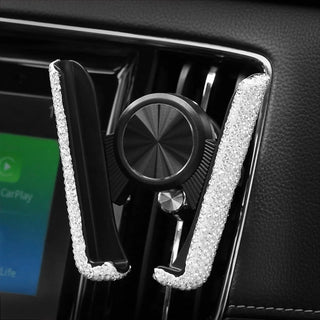 A Gabba Goods Bling Hands-Free Car Vent Mount is securely attached to a car's air vent, showcasing a sleek black body adorned with sparkling rhinestones along the sides. Situated behind the holder, a dashboard screen features a green interface. The car's luxurious black leather interior perfectly complements the bling design of the mount.